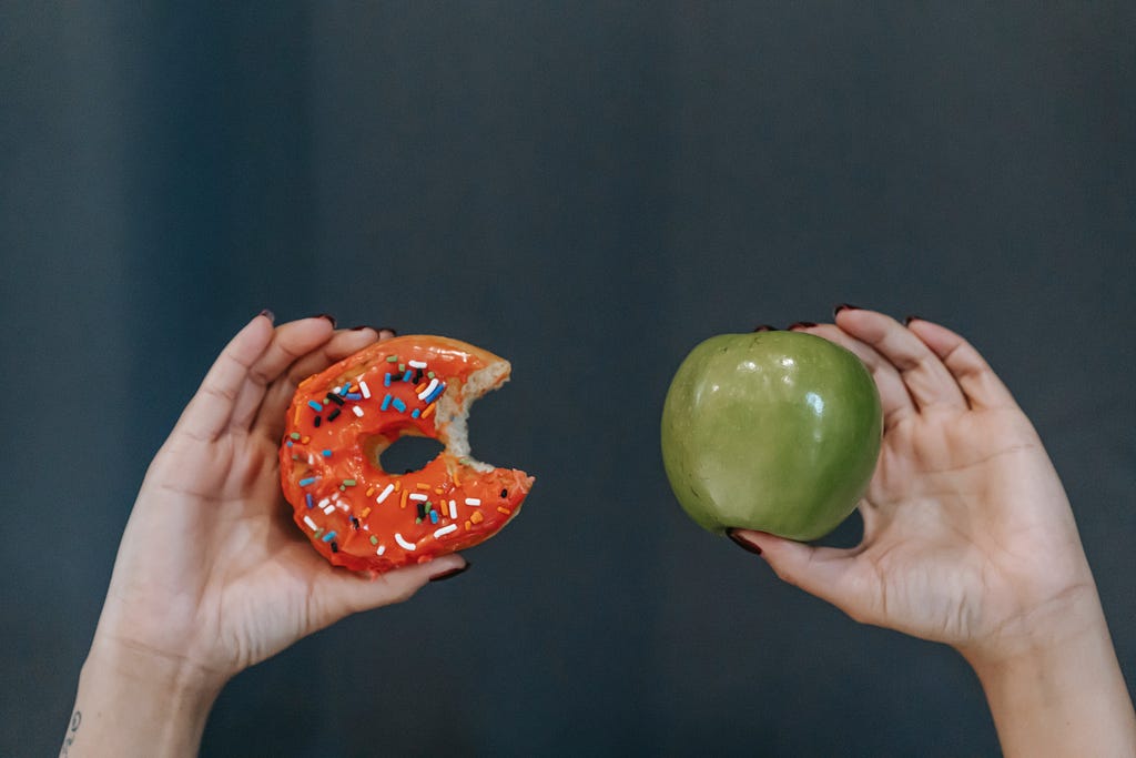 An image of two hands — one holds a half-eaten donut and the other holds an apple