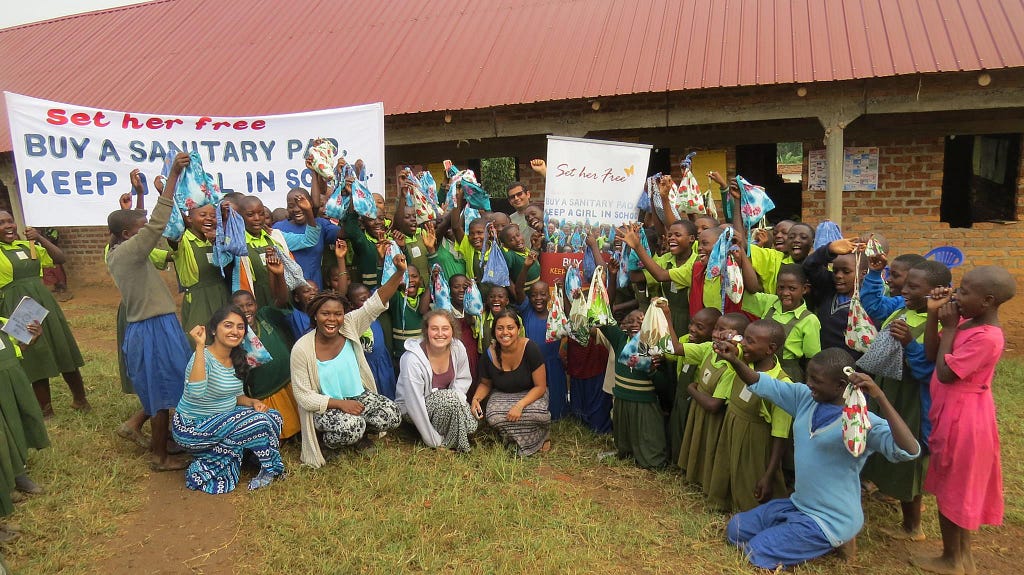 A large group of women and girls of all ages hold up bags of free sanitary pads in front of a house.