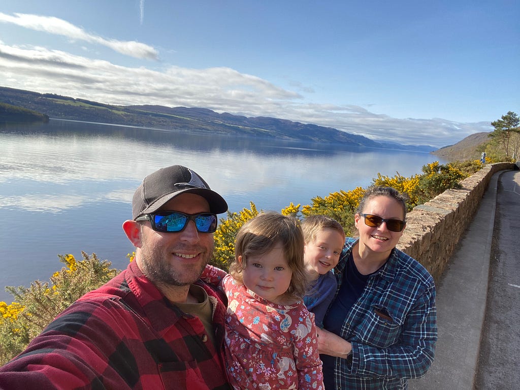 With my husband and two young children standing in front of a long rock wall in front of a large lake, with a clear blue sky.