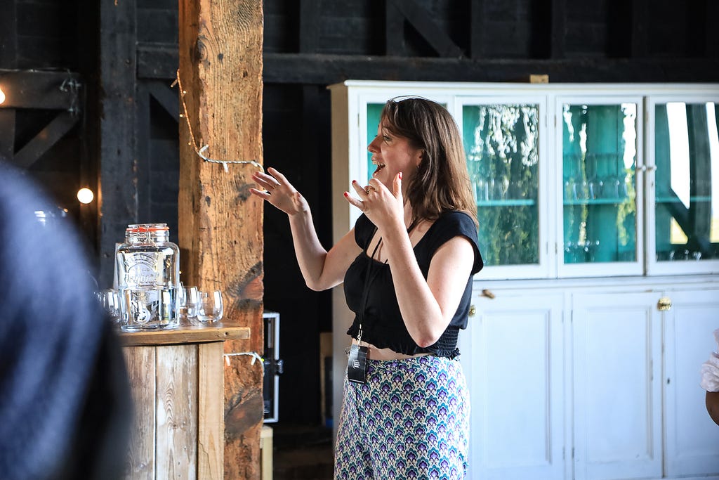 Black Design Guild team member Amelia Woods speaks to the group. Picture is of Amelia only with no on else in view. She is gesturing and speaking. She stands by a wooden beam and is wearing a black top and has a black and white print on her trousers.