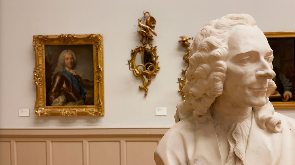 Photograph of a marble bust of a man looking to the right, with curly hair, on display in an art gallery. In the background, a gilded painting of a man in armor and an ornate wall sconce are visible on a white wall.