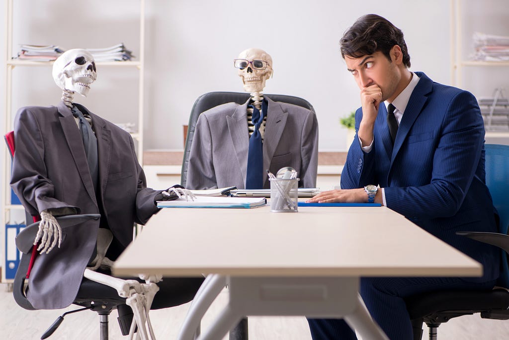 A man in a suit making a suspicious face having a business meeting with two skeletons wearing suits.