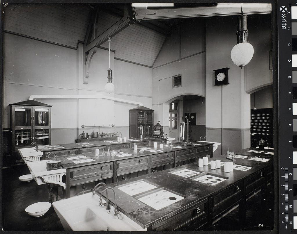 The interior of the dyeing lab in the main UMIST building on Sackville Street; Taken from the SW corner of the lab the shot is angled NE and shows three long wooden work tables in a single row each spanning nearly the entire width of the room. These all have porcelain sinks fixed to both ends by metal brackets.