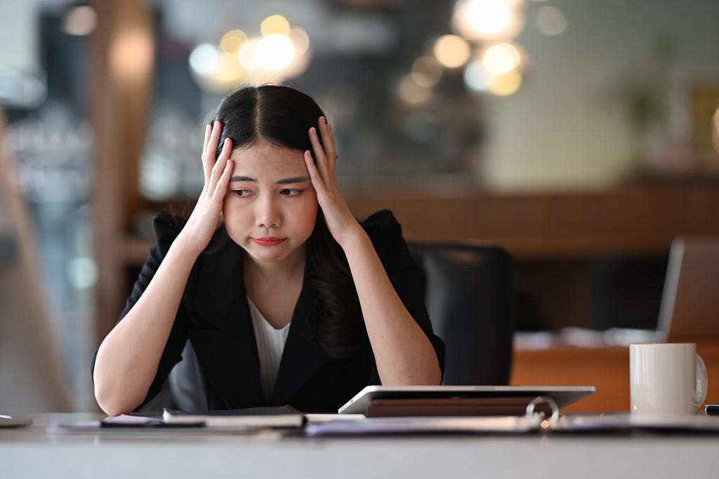 Frustrated female employee with hands on the temples of her head, sitting at a desk or table.