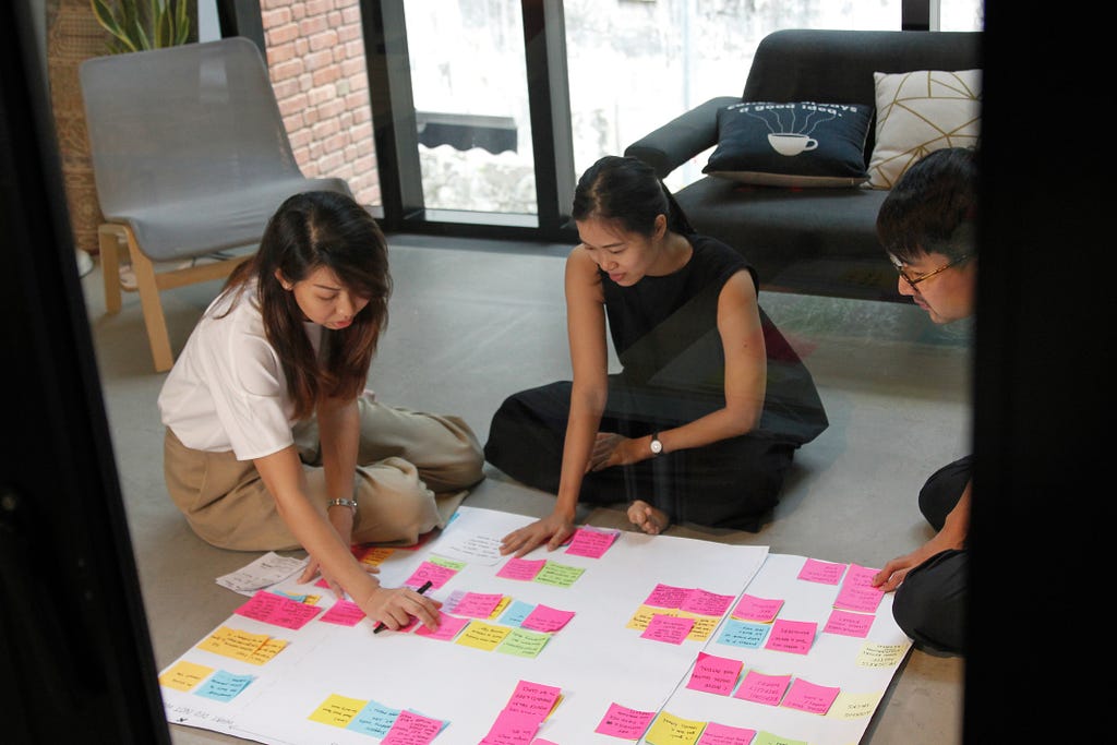 An image of three team members of 55 Minutes sitting on the floor, having a discussion around the post-it notes pasted on a big board placed on the floor.