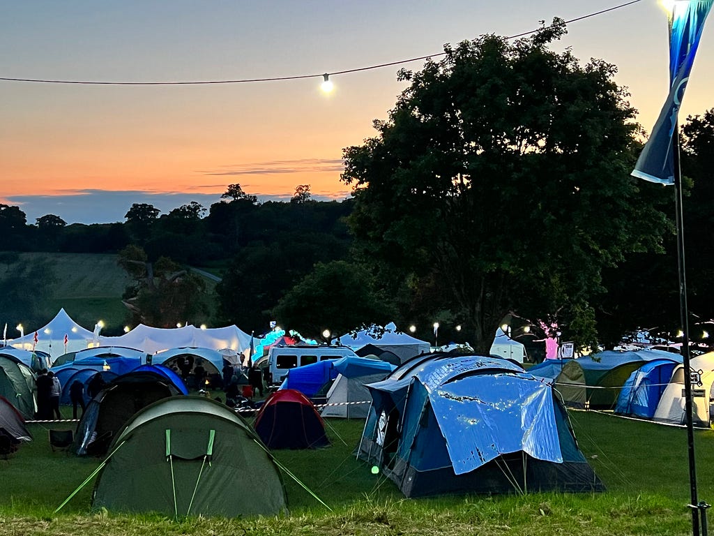 Tents in a field at dusk. A lightbulb glows on a cable above them, against the orange sky.