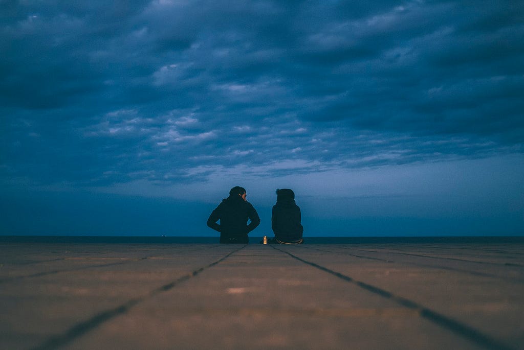 Two people having a private conversation sitting on something like a boardwalk at dusk with clouds overhead and the horizon stretching out before them.