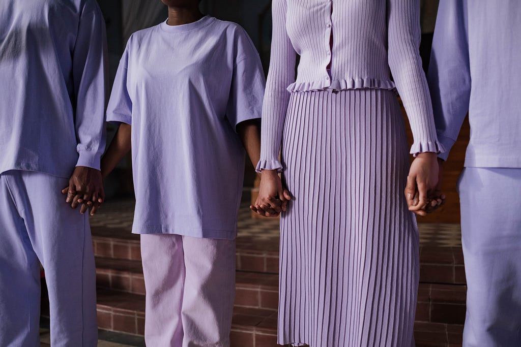 4 persons holding hands, all wearing lavender monochrome outfits; the taller woman stands out with a ruffled top and pleated skirt