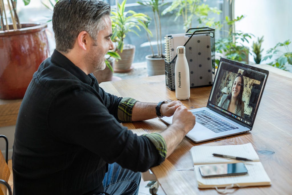 A man sitting at his desk, conducting an online interview on a laptop. Laptop screen shows a woman that he is talking to.