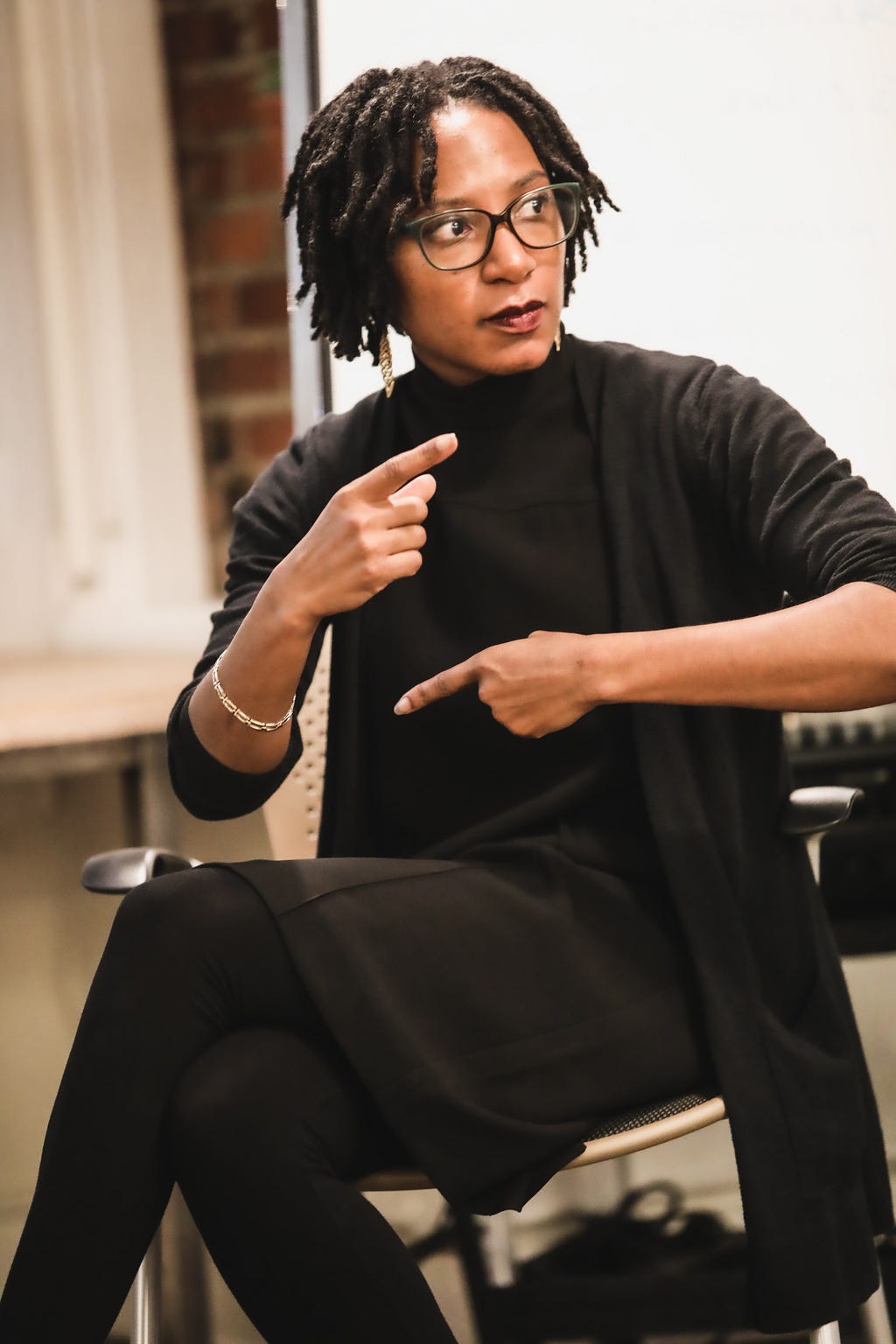 Lindsey (the article author) sits in a chair wearing a chic all-black outfit. Her index fingers point in opposite directions across her body as she wears a focused expression.