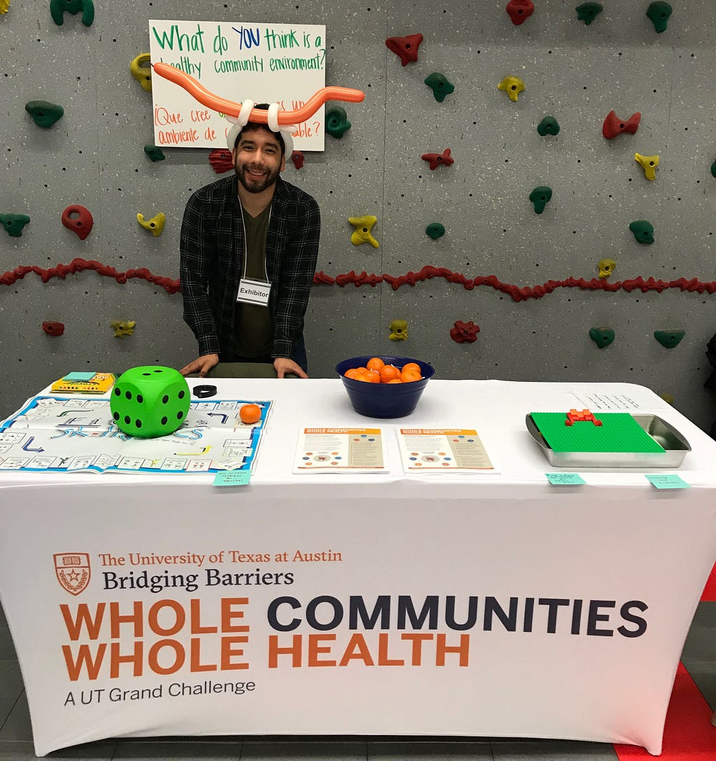 A UT Austin graduate student wears a longhorn hat and stands beside a Whole Communities–Whole Health table at an event.