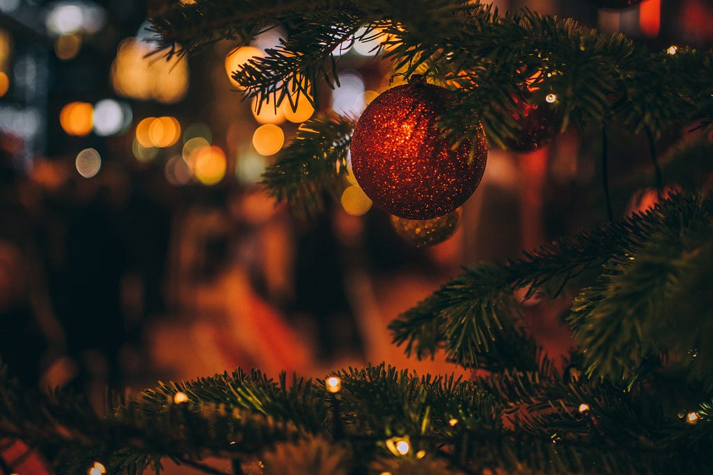 Image is of a close up of a red Christmas bauble hanging on a Christmas tree. Christmas lights are in the background