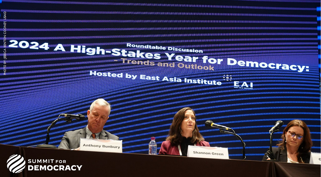 Three people each sit behind a microphone at a long table during a public discussion. A projection on the wall behind them displays the topic of their discussion: “2024 A High-Stakes Year for Democracy.”