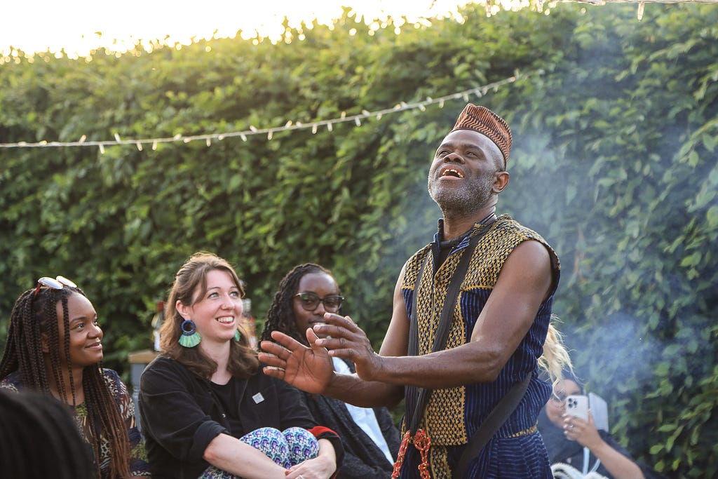 Storyteller Usifu Jalloh, wearing a brown hat, brown and blue top and blue trousers has his hands out and face turned upwards. He has a drum strap around his neck. He’s surrounded by guests looking at him. Beghind him is a green bush and above hium is a string of fairy lights.