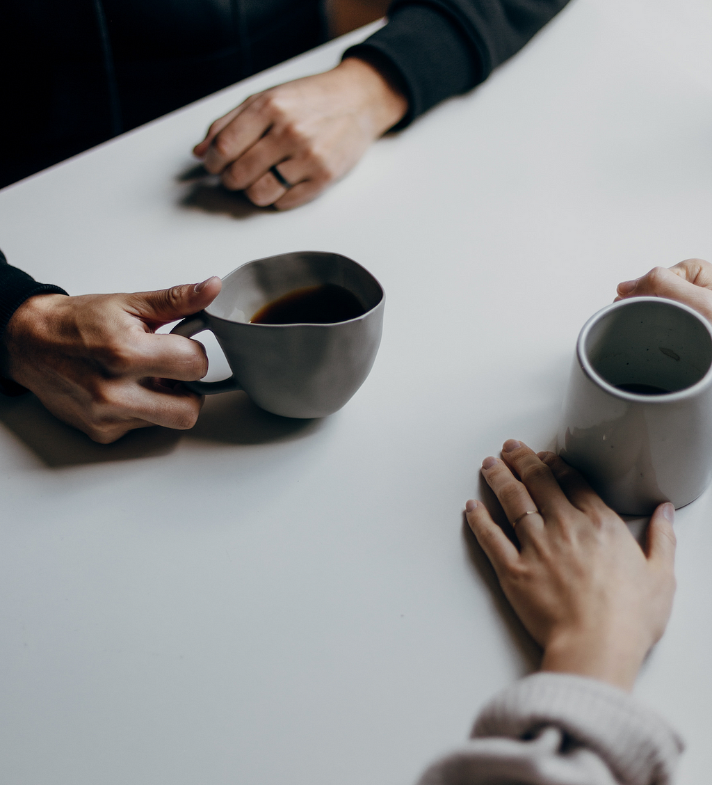 A guy and girl sitting face-to-face over a table, each holding a mug of a hot beverage. Both are seen wearing a wedding ring, indicating it’s a couple conversing.