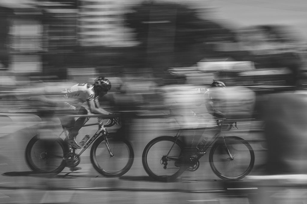 Black and white image of two cyclists riding in a city with extreme motion blur.
