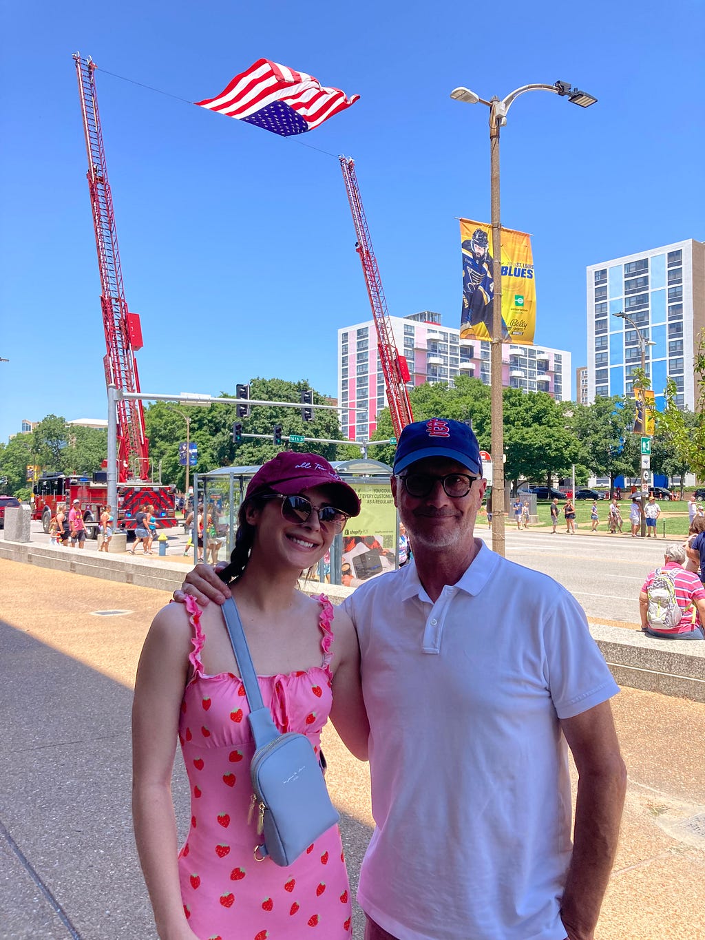 Colorful photo of the writer with his younger daughter at Pride parade with American flag flying, between extended ladders of two fire trucks, in a blue sky behind them. Photo credit is to “a fellow citizen.”