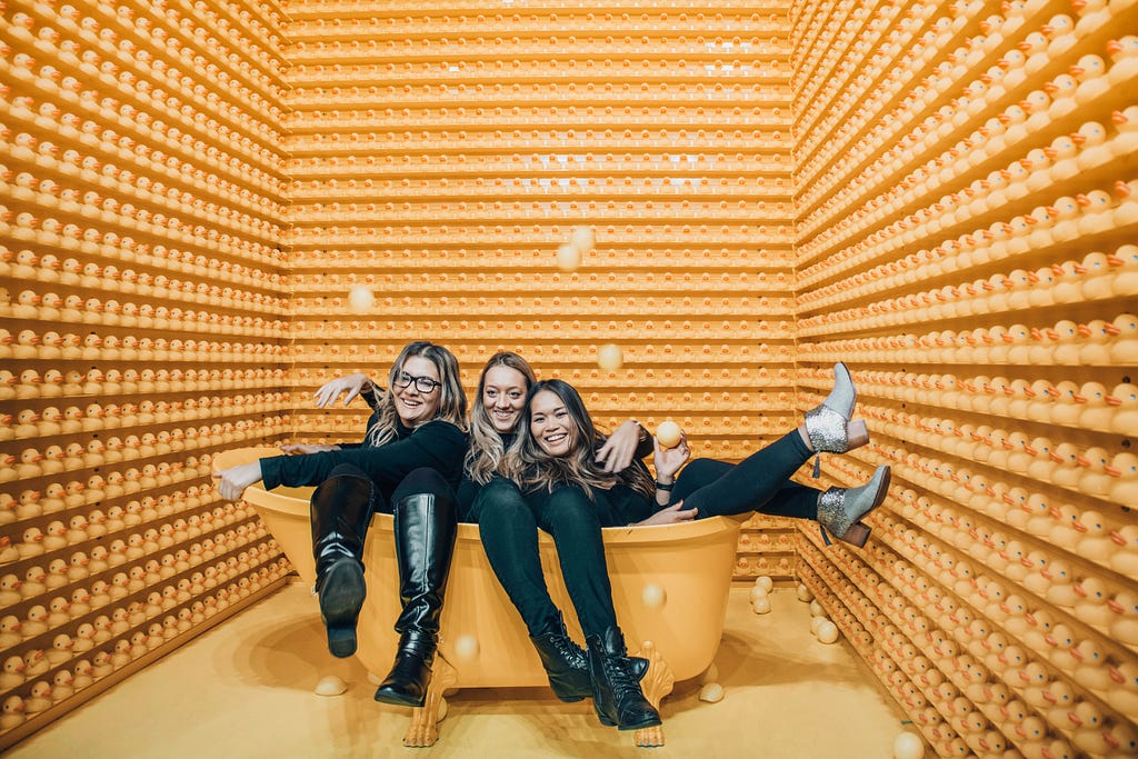 three women sitting in a yellow bathtub (fully clothed) smiling with a yellow backdrop