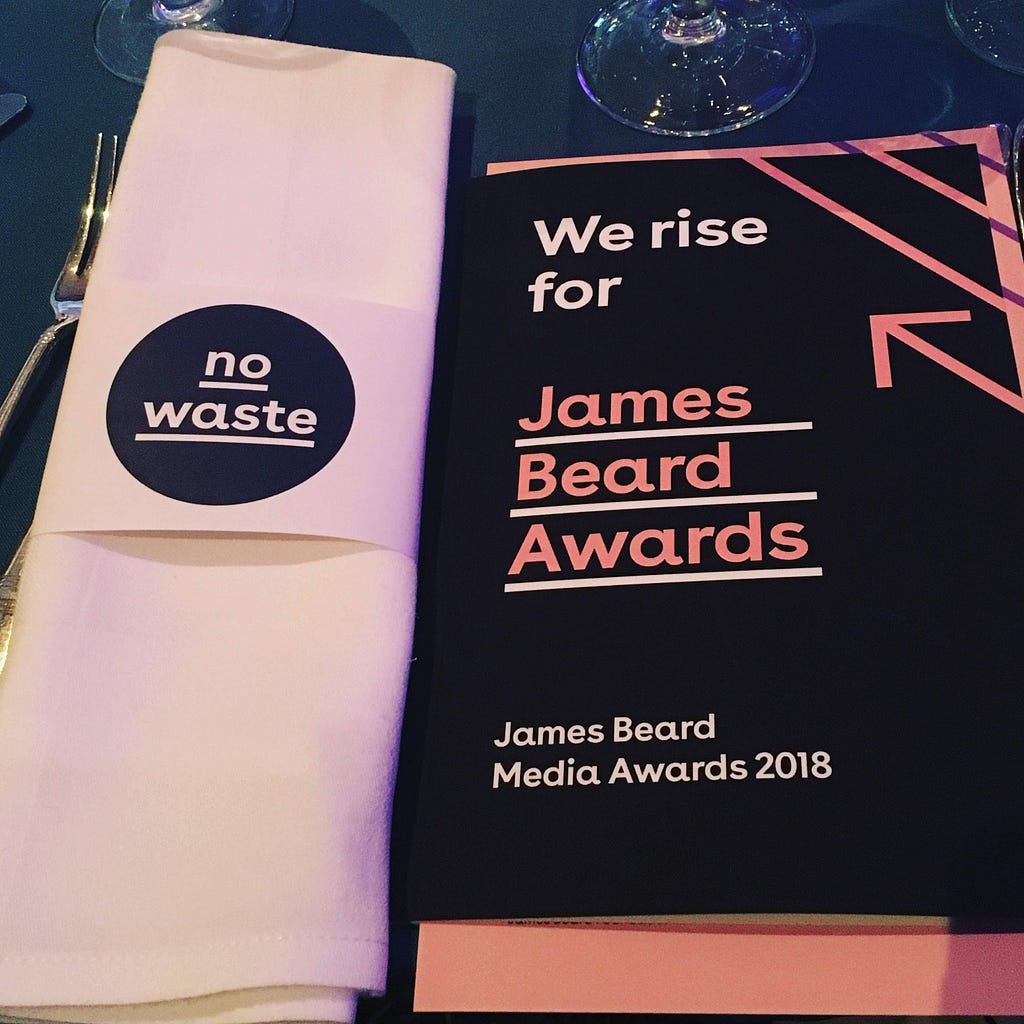 On the left, a linen napkin wrapped in a holder that says “no waste” on the right is a black printed program with pink writing that says “We rise for” James Beard Awards, James Beard Media Awards 2018.