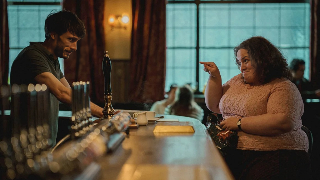 Left: A bartender leans onto the bar. Right: Across him, a woman points at him as she holds back a smile as if in the middle of telling a joke