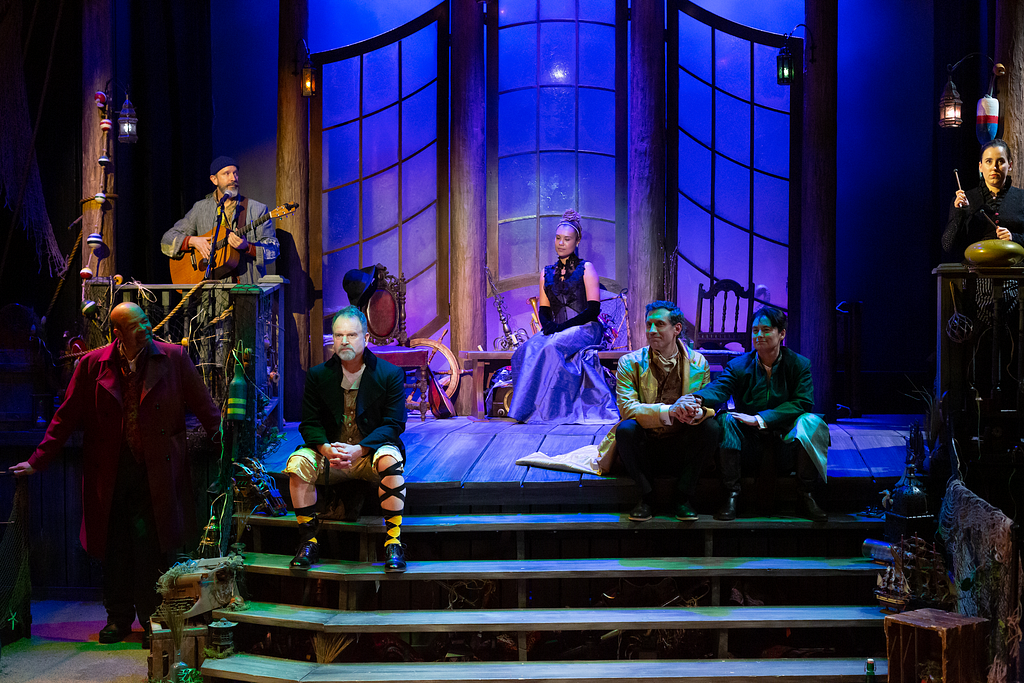 A group of actors sits about a stage in blue and purple light while another actor plays guitar and sings into a microphone.