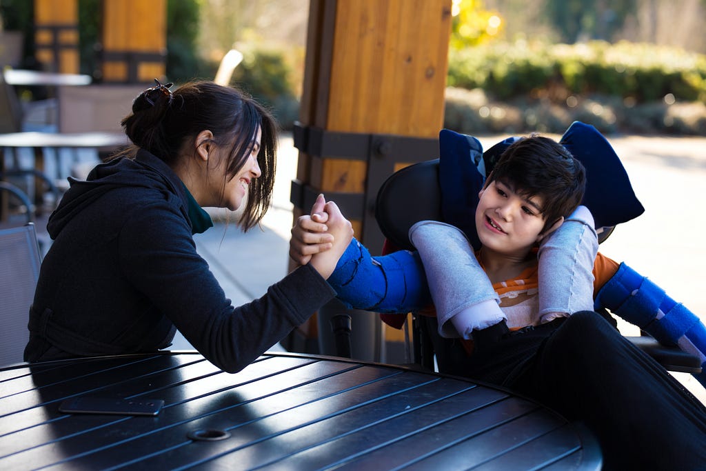 An adult and a child in a wheelchair, sitting at a table outside, and arm wrestling with smiles on their faces