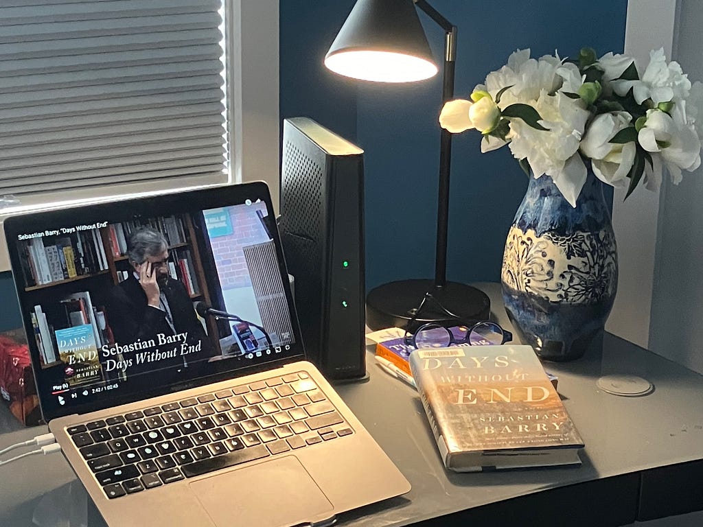 Sebastian Barry video plays on a laptop on a desk with a print copy of the book and a vase of flowers