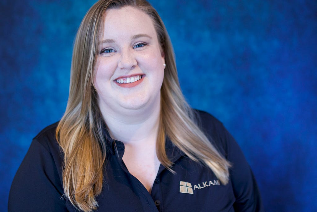 Claire Tolbert stands in front of a blue background wearing an Alkami logo polo