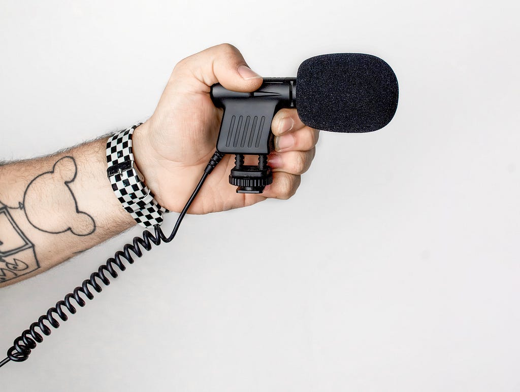 Mão segurando um objeto que parece uma arma mas que, na verdade, é um microfone apontado para frente.