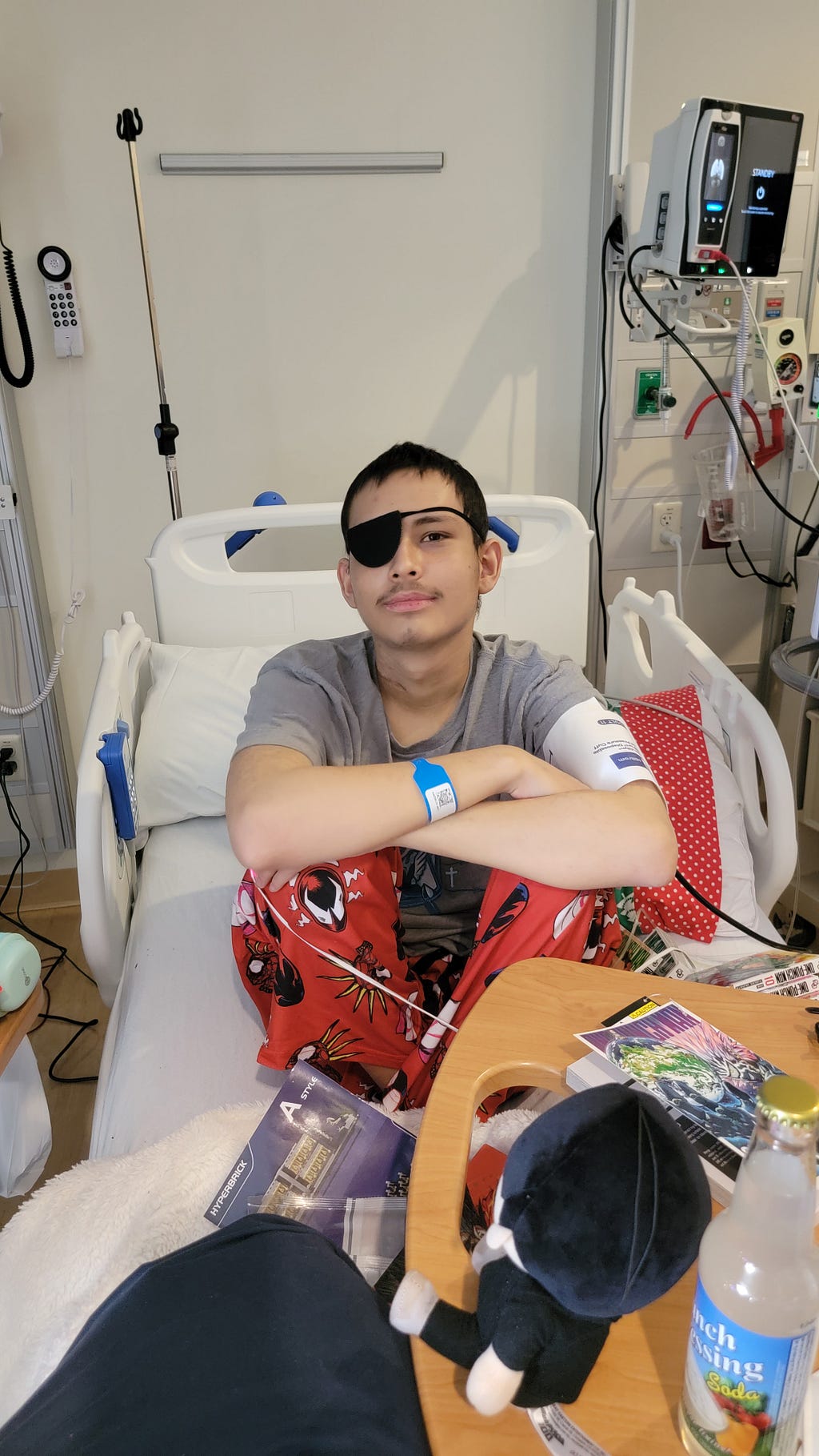 A young man in a hospital bed wears a black eye patch, sitting cross-legged with a calm expression.