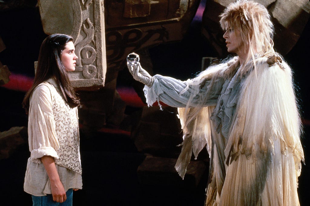 A scene from the movie, Labyrinth: Sarah, a young, white female in poet blouse, vest, and jeans, stands to the left facing the Goblin King played by David Bowie. In the background, broken pieces of the labyrinth float in space. The Goblin King has a feathered classic rock mullet. He wears a white poet’s blouse and a white cape with brown accents that is reminiscent of feathers. The Goblin King holds out a clear, shiny sphere for contact juggling. Sarah stares at it transfixed.