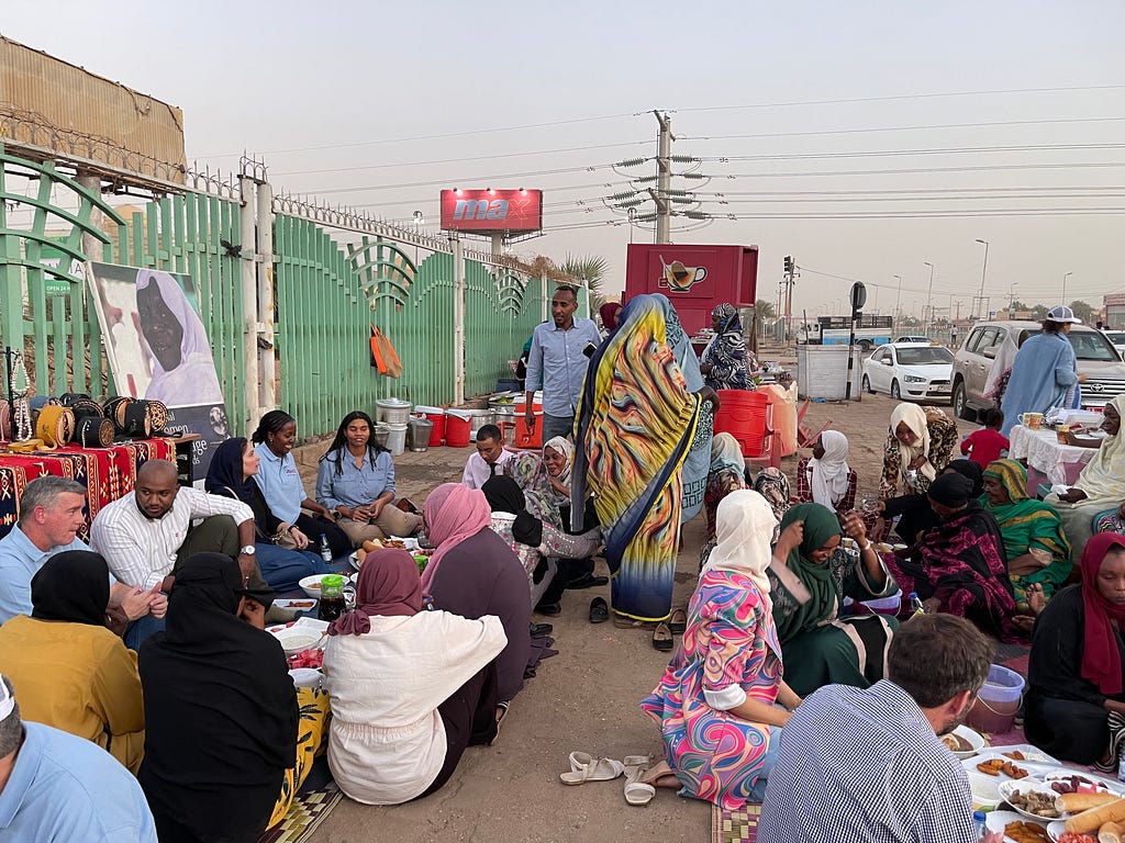 A group of people partake in an iftar, or evening meal, while sitting on the ground on the side of a roadway.