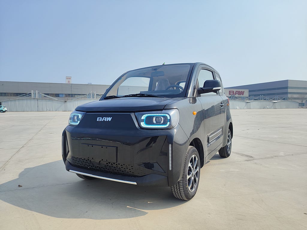 Un microcoche de color negro con luces LED. Está aparcado en una zona industrial con fábricas al fondo.