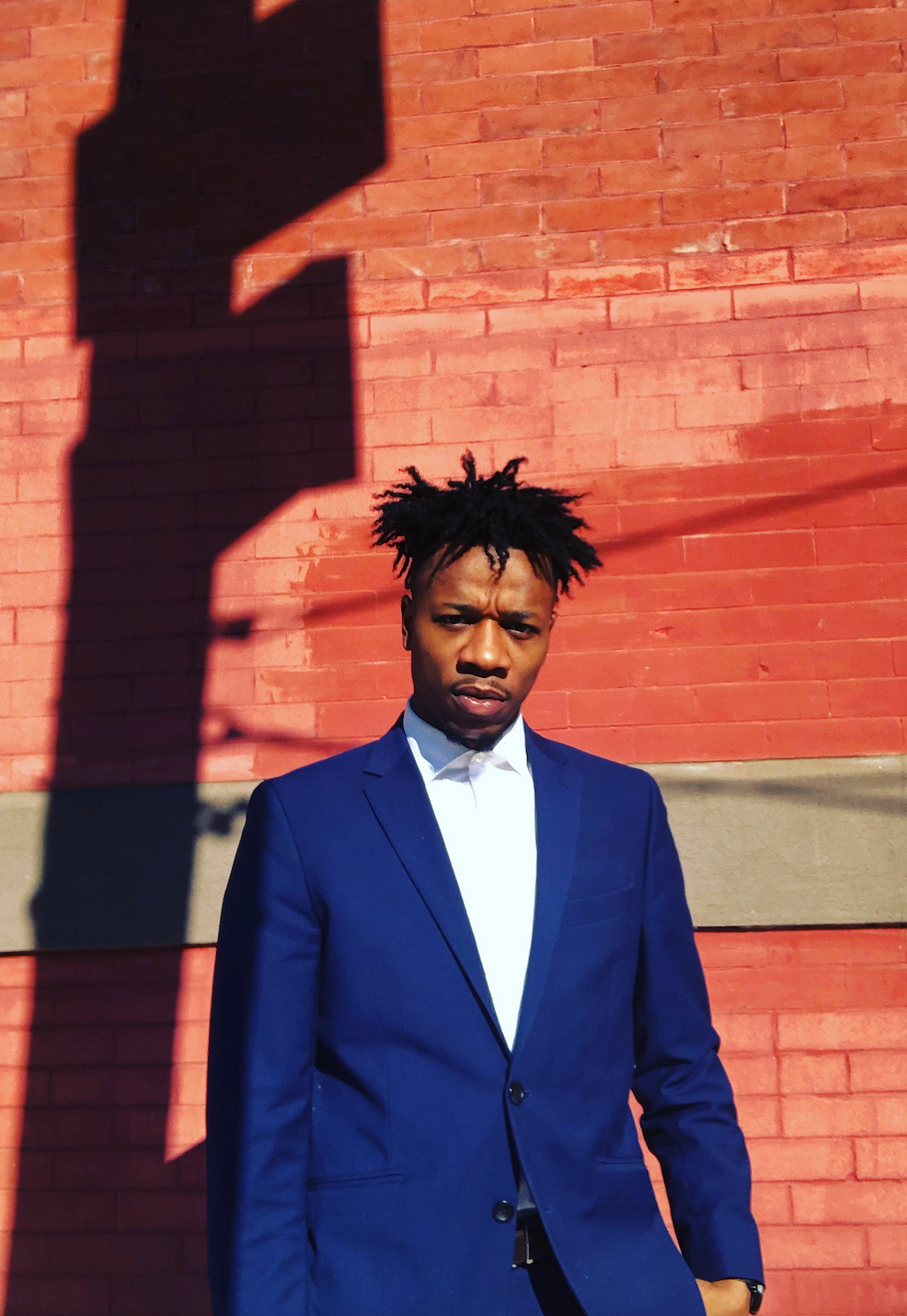 Director Tyree Barnes is photographed here in a white button down shirt and navy blue blazer standing in front of a reddish brick wall.