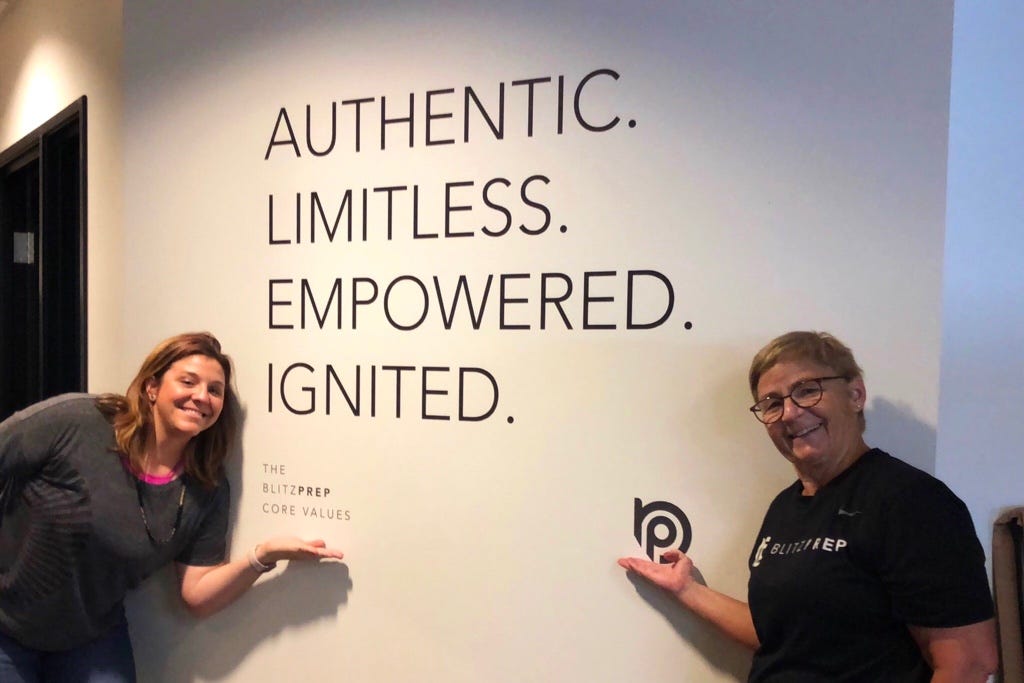 Two women smile at the camera while pointing to a banner that reads “Authentic, Limitless, Empowered, Ignited.”