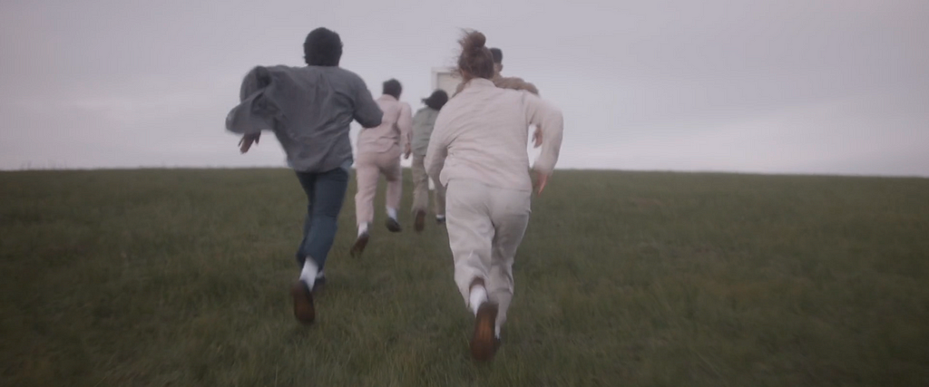 Members of the Project Home Ensemble run towards a white door in the middle of a grassy field.