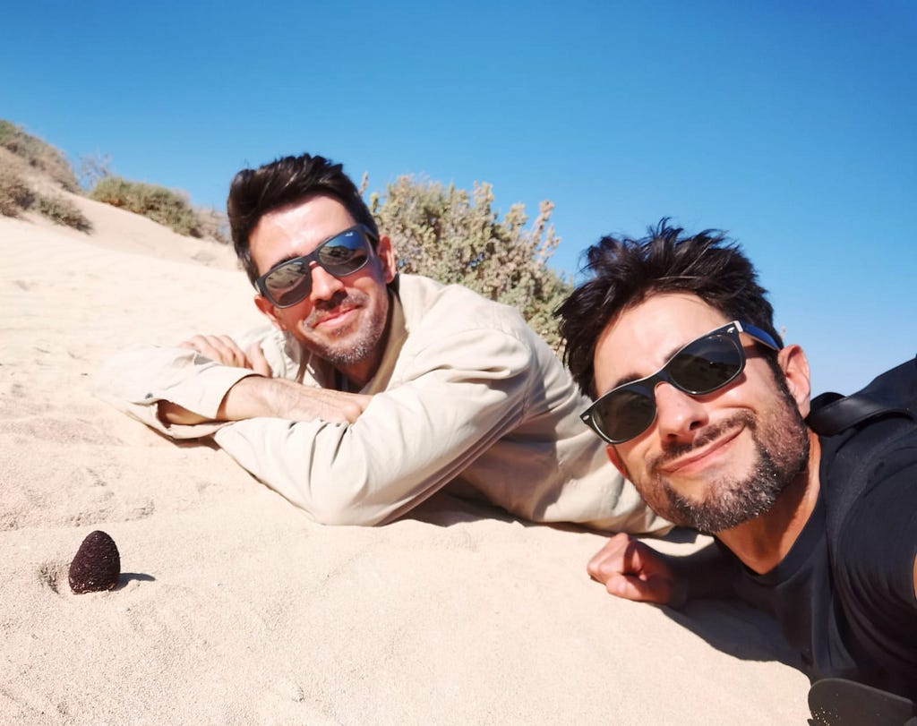 Matias and Chris smiling and laying in the sand next to a Cynomorium coccineum plant jutting out of the sand.