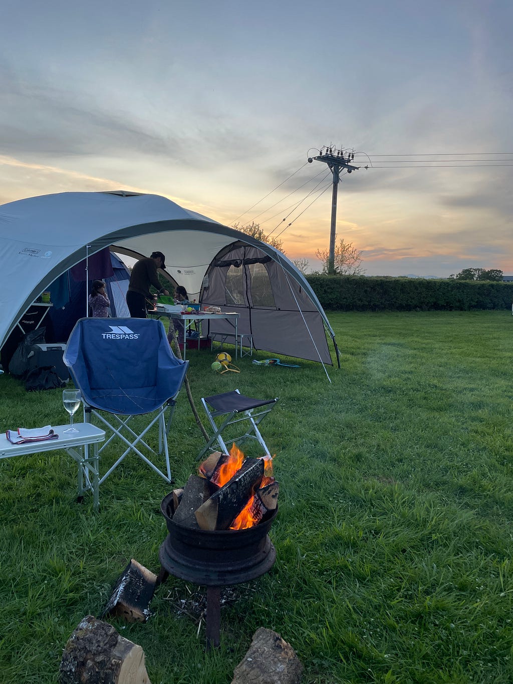 Image shows a gazebo in a campsite field at dusk, with a campfire and campign chair in the foreground. A bench next to the campign chair has a glass of wine on it.