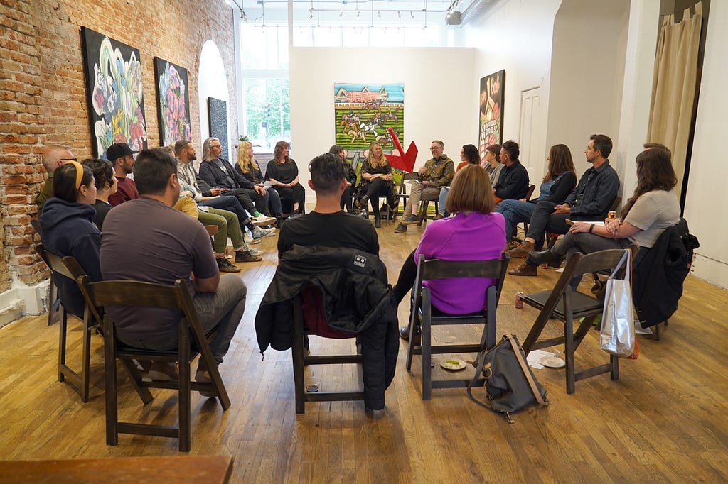 A group of 30 people sit in a circle of black chairs. The photo is taken from behind the group. They sit in an art gallery surrounded by brightly colored paintings.