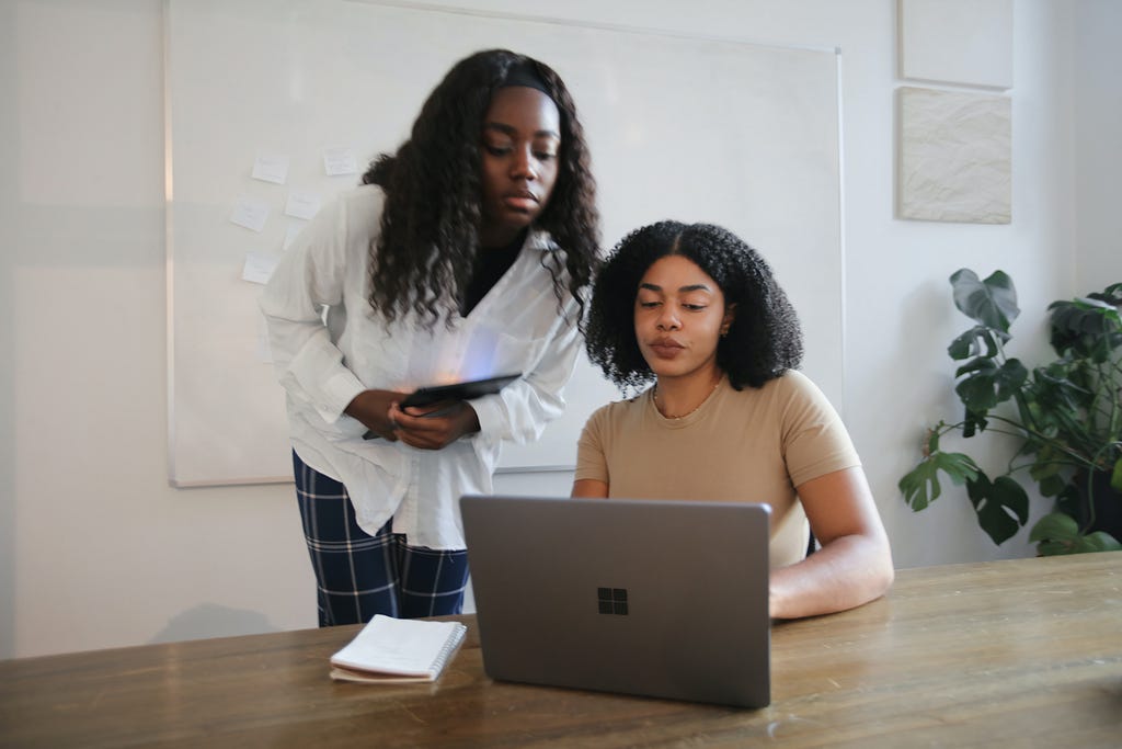 Two African American young women look at a laptop in an office with white walls.