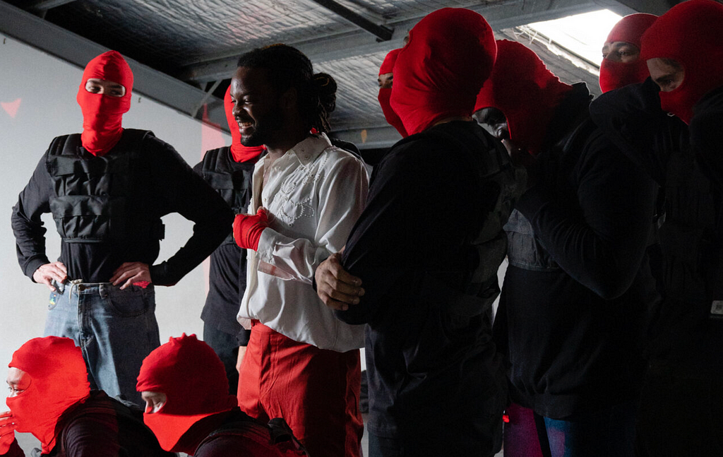 Genesis Owusu is surrounded by his ‘goon squad’ during a behind the scenes moment preparing for his 2021 ARIAs performance. Both he and his dancers/squad are dressed in solid black, red and white colours with his dancers also wearing red balaclavas. Genesis is smiling and looking off camera.