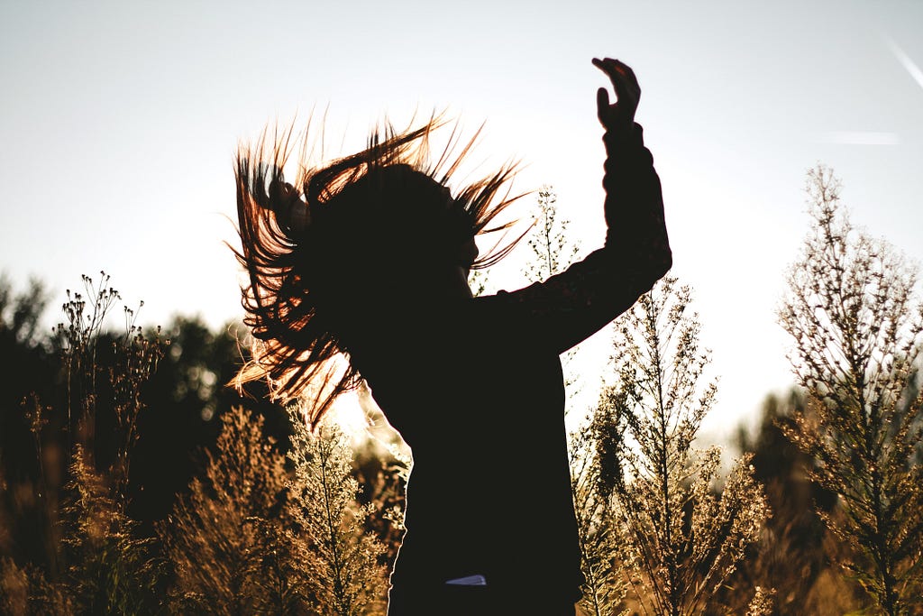 A person with long hair is in a silhouette in a meadow and they are moving their body in what appears to be an expression of joy and freedom.