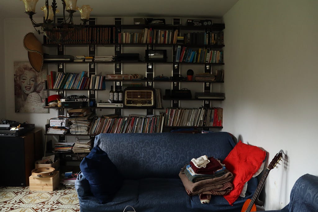 A cozy interior scene with a well-worn couch, folded blankets, and a guitar resting nearby, all framed by wall-mounted shelves filled with books, records, and various vintage objects.