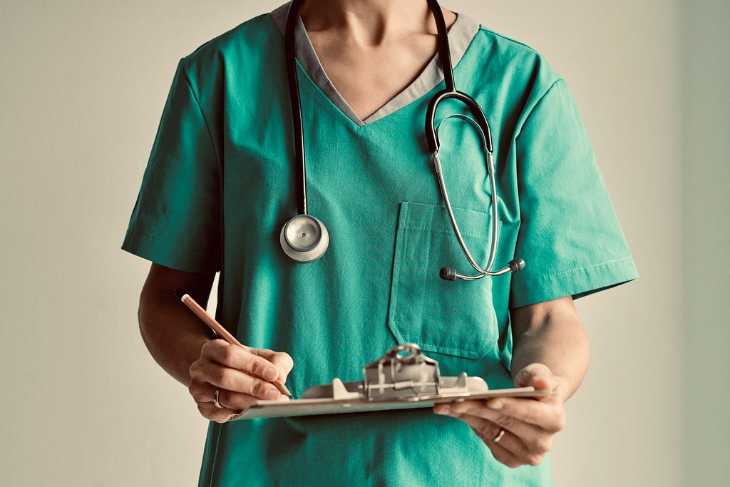 A young woman in green hospital scrubs and stethoscope holding a clipboard and pen