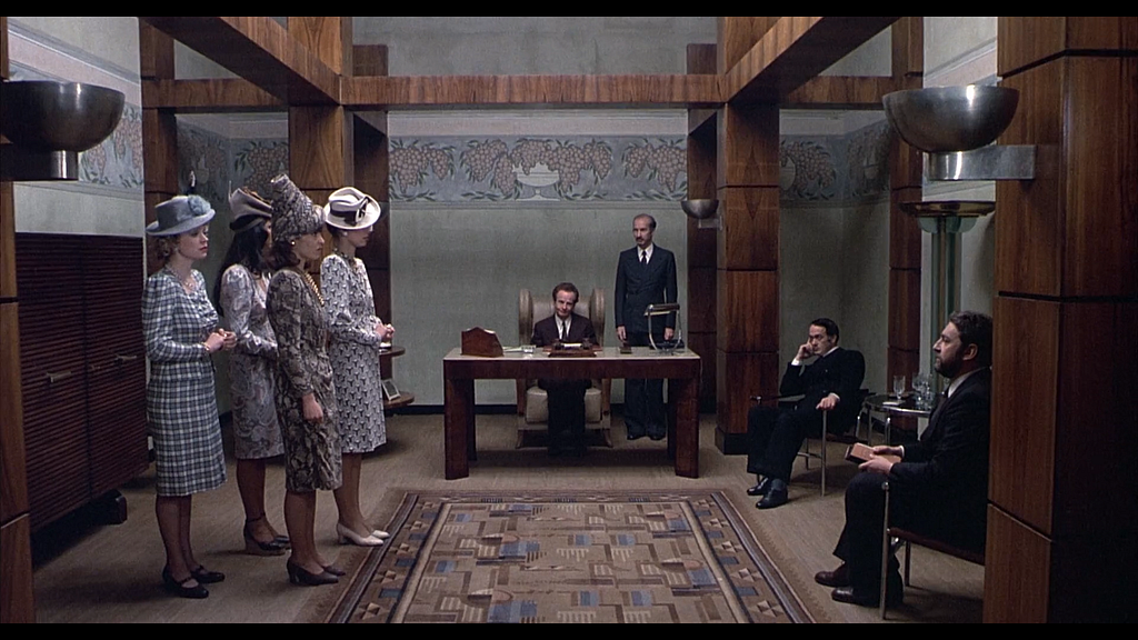 A screenshot from the film: we as the viewer are looking into a room in which the four daughters (well-dressed, in light colours) are gathered together in the left and the four libertines (all in black suits) are sat down in the centre and to the right. The body language of the daughters is tense and afraid; the libertines are relaxed and a little sinister.