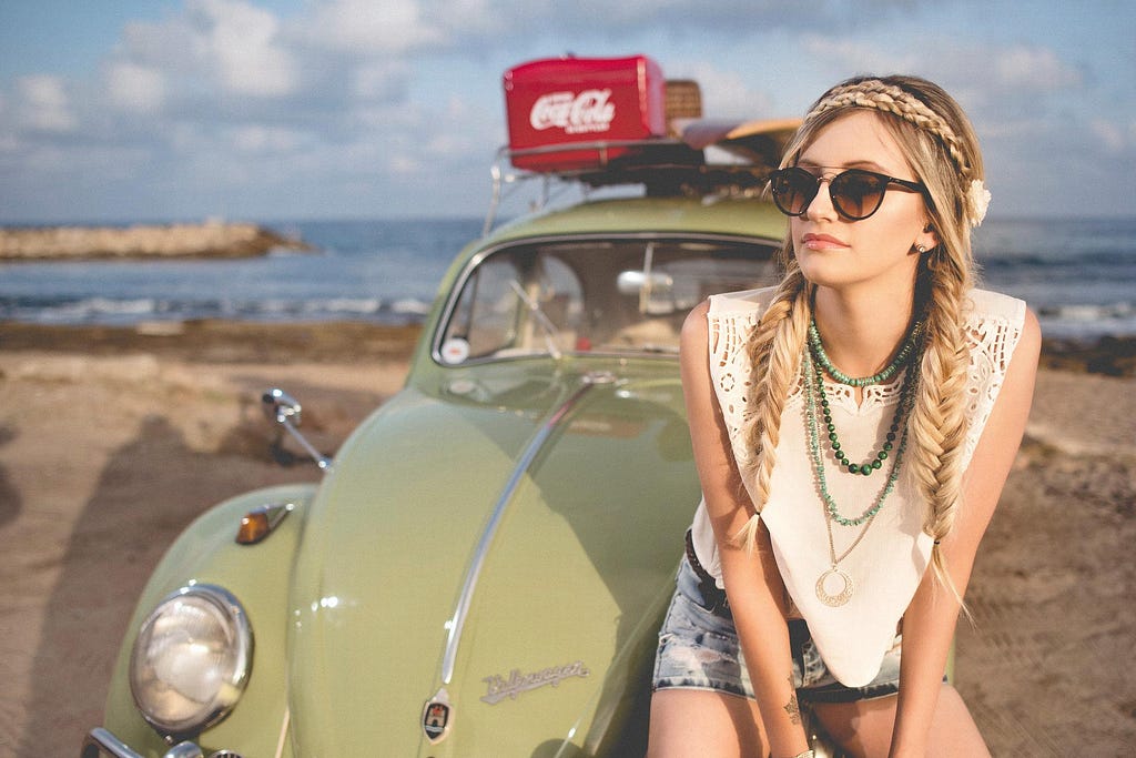 A young women looking like a hippy, sits on the front of her VW beatle, looking off camera.