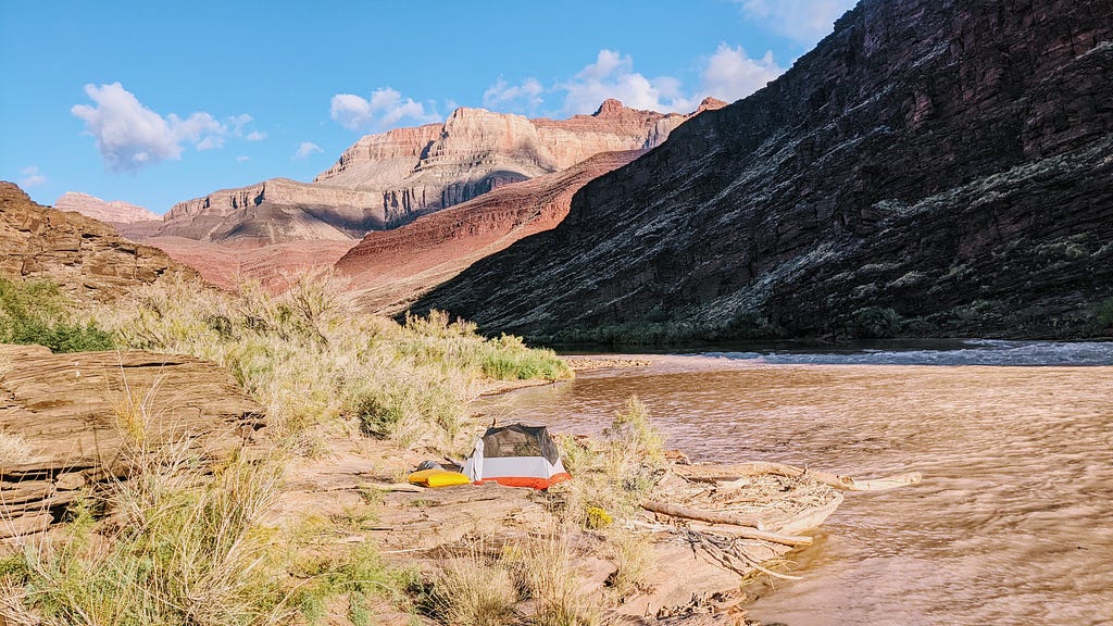 A camping tent set up on the banks of the Colorado River in the Grand Canyon, with the rim of the canyon visible in the distance