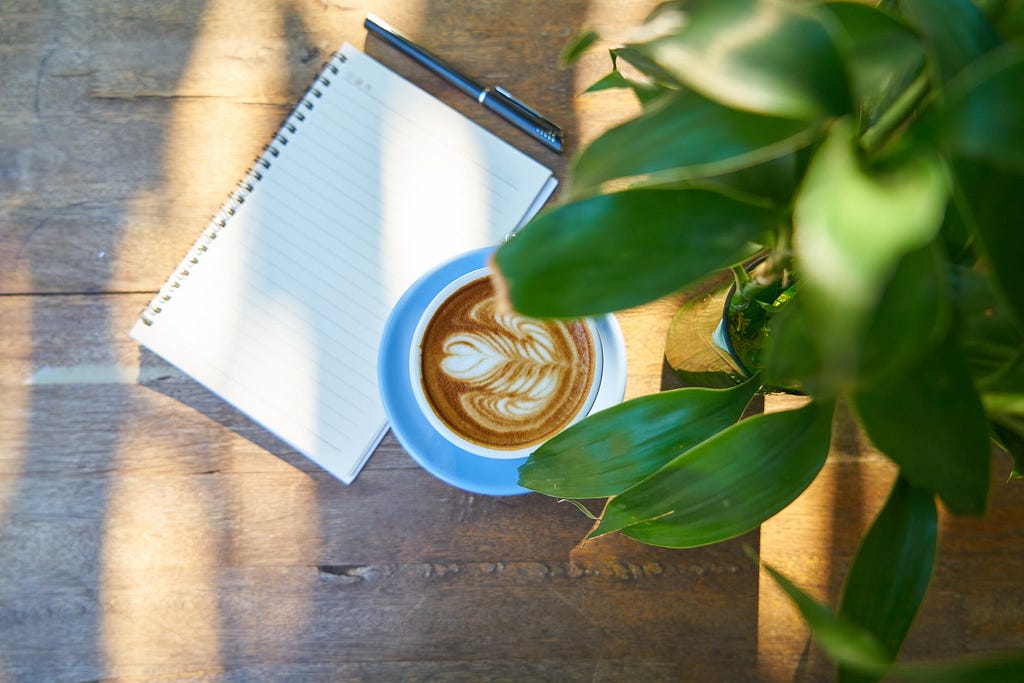 Imagem encontrada no site Pexels, nela podemos ver uma superfície marrom que remete a madeira, sobre ela há um bloco de notas branco, com uma caneta preta ao lado e uma xícara branca com café. Em primeiro plano da imagem há uma planta que sobrepõem os outros objetos da foto.