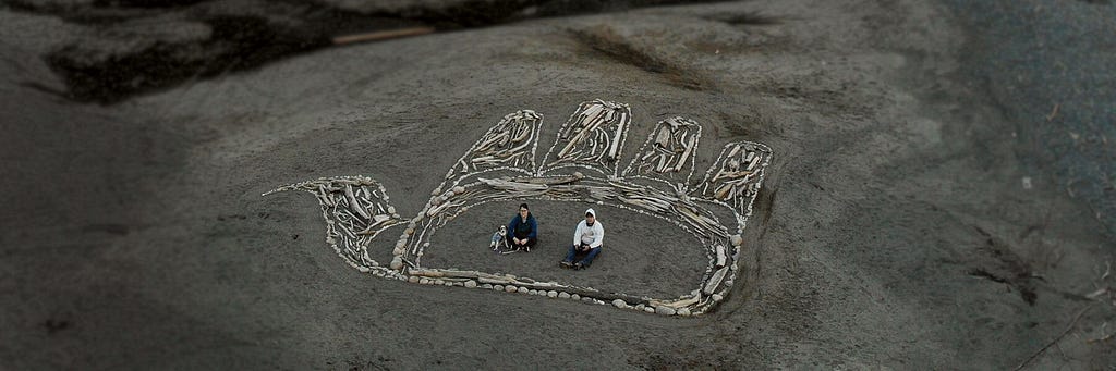 A massive Gitxsan formline artwork of an open hand made of driftwood and stones on a sandy riverbank. Two people and a dog sit inside the palm of the hand.