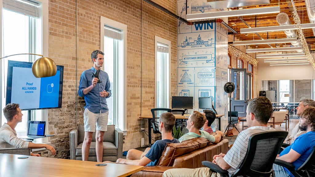 A casually dressed male speaker talking in front of a few people in what looks to be a startup office